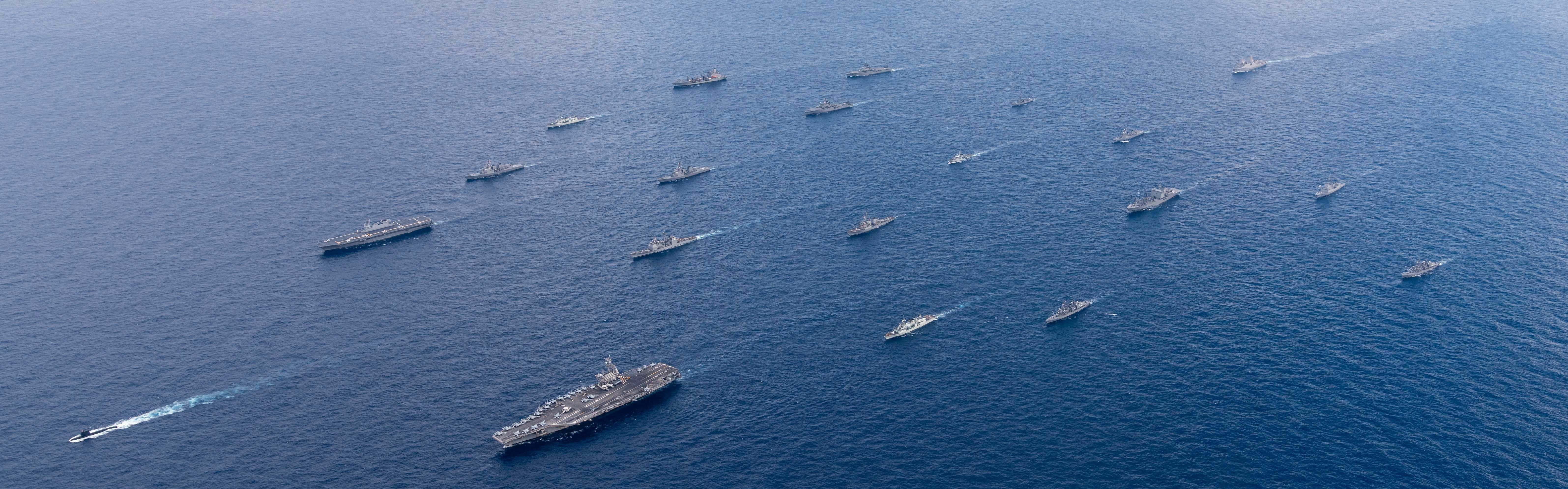 Gray naval military vessels from the US, Australia, Japan, and South Korea sail in formation on a deep blue ocean. The sky is mostly clear and blue with a few thin clouds just on the horizon.