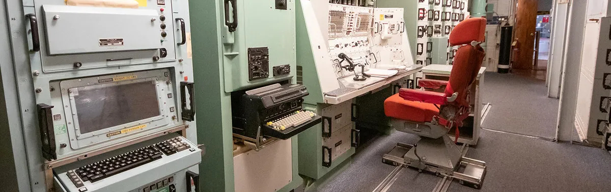 Inside a launch control room for the Minuteman missiles, a wall of aged technical applications includes old keyboards, bright red warning lights, and a landline telephone. A red chair sits bolted to the floor in front of the control panel. 