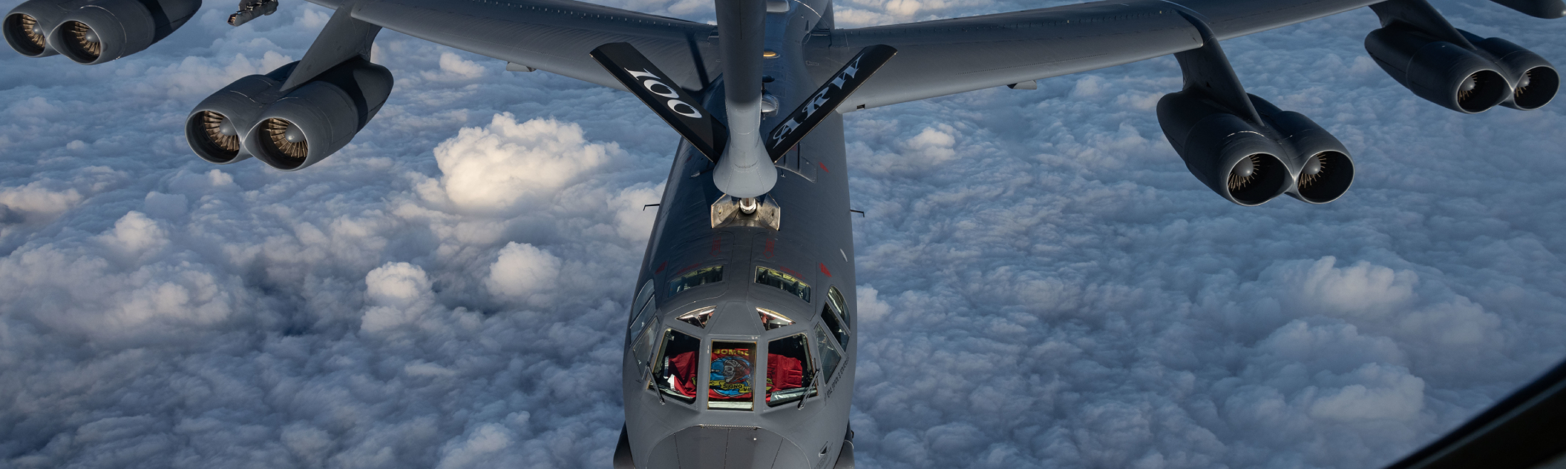 A gray aircraft is shown in flight over fluffy white clouds. White letters are painted on the plane indicating it is from the 100th air refueling wing. 